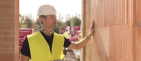 Builder on construction site touching clay block wall, construction workers and clay block pallets in the background