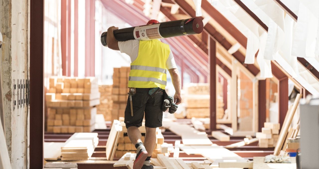 Urban roofer carrying underlay sheet on his shoulder in renovated attic wearing hard hat and safety jacket