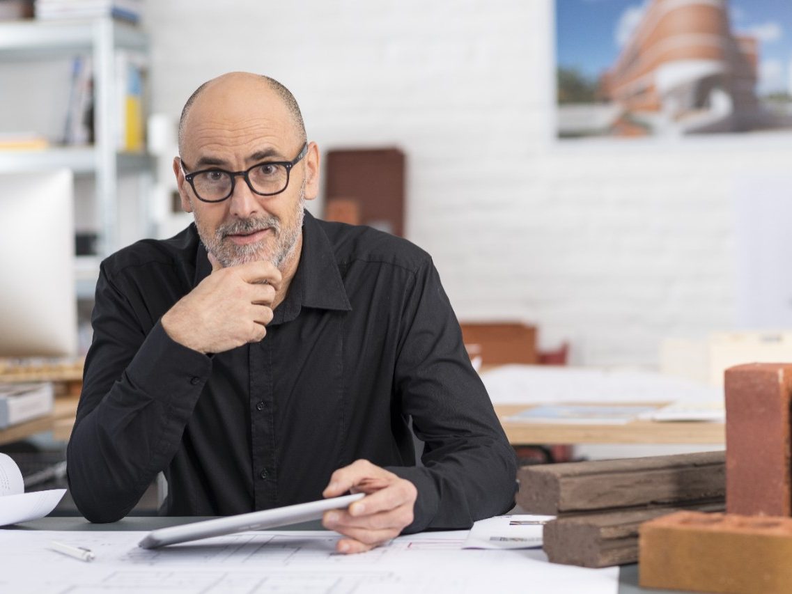 Senior architect sitting at office desk with blueprint holding tablet facing camera