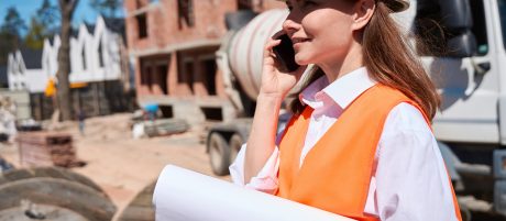 Qualified woman architect in hardhat and safety vest talking phone, receiving instructions from the general contractor, spending time on construction site