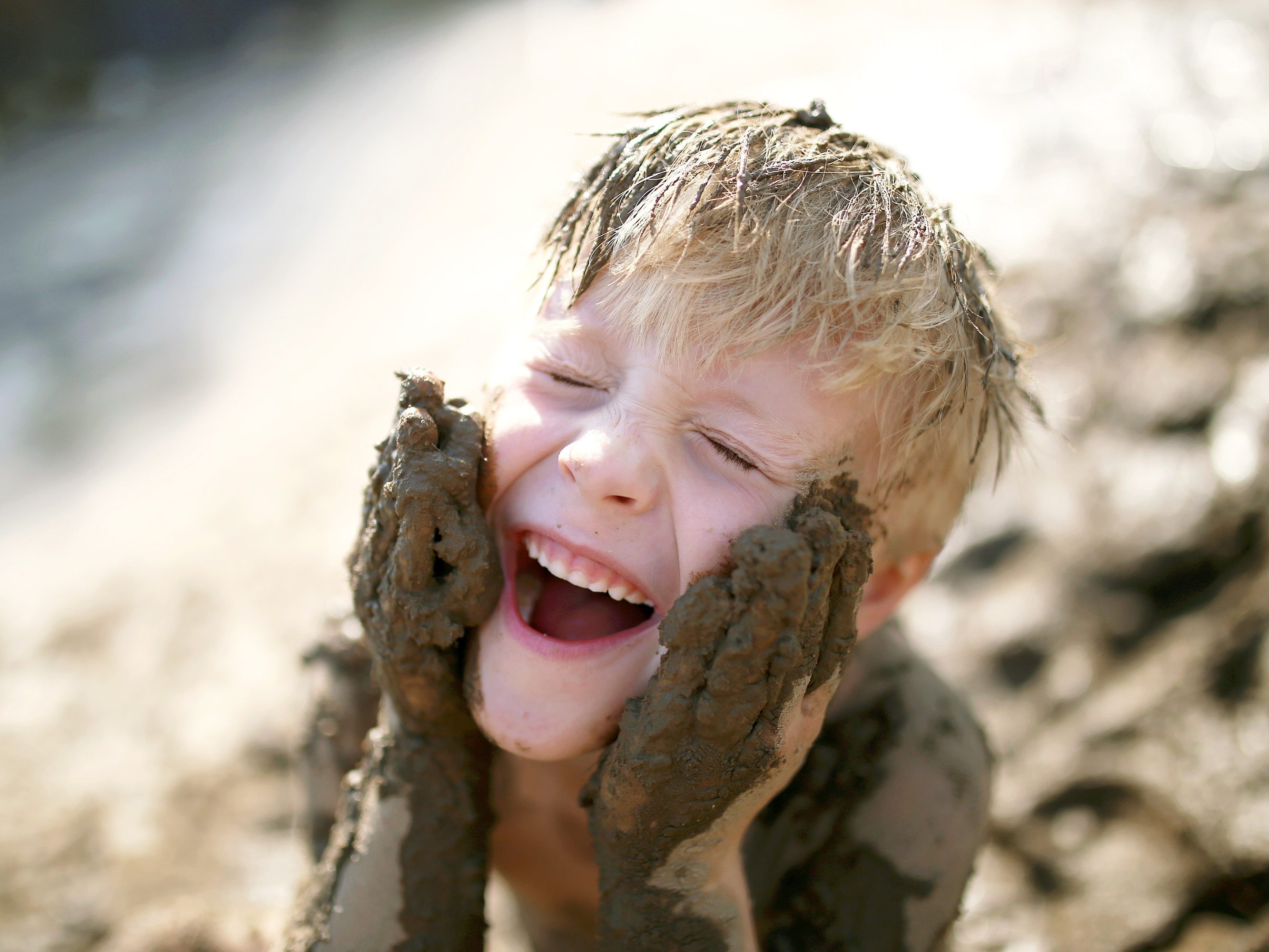 A cute little boy child is laughing as he plays outside in the mud and rubs dirt on his face with his hands.