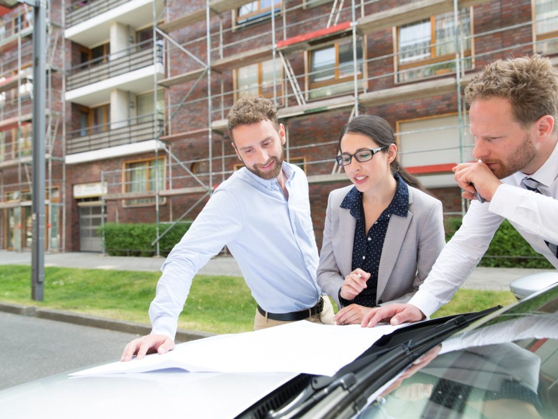 Architects or planners looking at a blueprint placed on a car bonnet, in front of a building which has a reddish brick façade and is covered in scaffolding