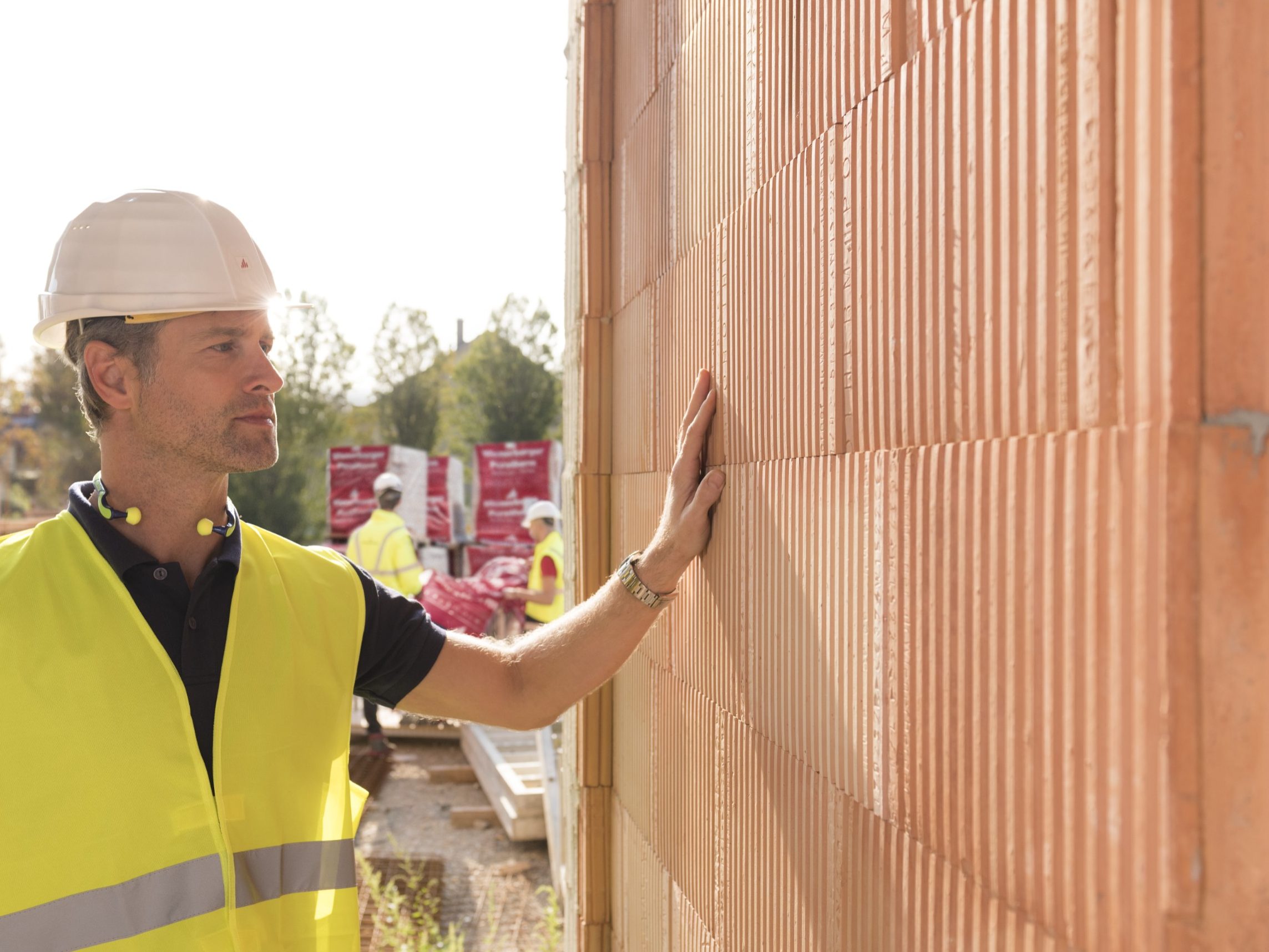 Builder on construction site touching clay block wall, construction workers and clay block pallets in the background