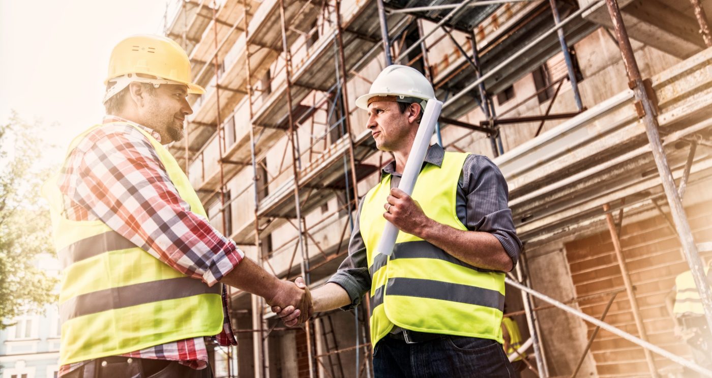 Handshake of two man wearing reflective clothing and work helmets o a construction site, scaffolding in background.