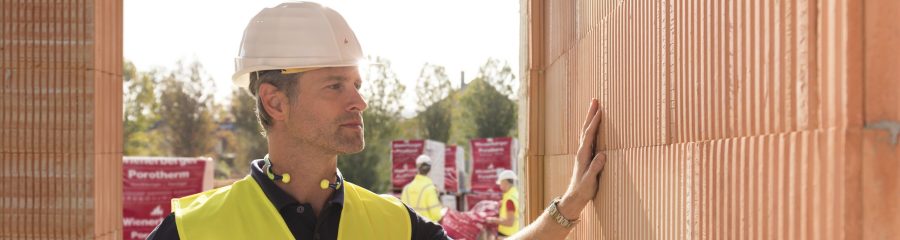 Builder on construction site touching clay block wall, construction workers and clay block pallets in the background